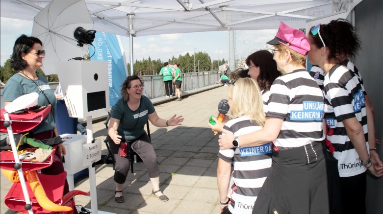 Gruppierung von sechs Personen in gestreiften Shirts, die auf eine Frau in einem blauen Shirt reagieren. Im Hintergrund ein Zelt mit einem Fotostand.